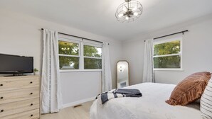A bedroom with a bed, a dresser, a TV, a mirror, and two windows with curtains. - Charming 4BR near Convention Ctr., Main St, & LU -just 11 minutes to STL Airport (Saint Charles)
