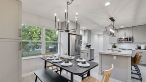 A modern kitchen with a central island, stainless steel appliances, and a dining area with a black table and chairs. - Charming 4BR near Convention Ctr., Main St, & LU -just 11 minutes to STL Airport (Saint Charles)