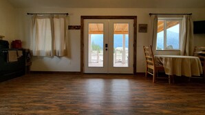 A room with a wooden floor, a table with a tablecloth, a chair, a sofa, a TV, and a view of the outdoors through a door.
