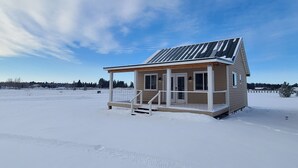 A small, single-story cabin with a covered porch and solar panels on the roof, surrounded by a snowy landscape.