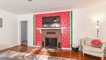 A living room with a fireplace, a patterned rug, a sofa with a watermelon pillow, and a wall with a watermelon design.