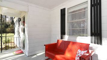 A red bench on a porch with a white exterior wall, a window with black shutters, and a door leading to a covered area.