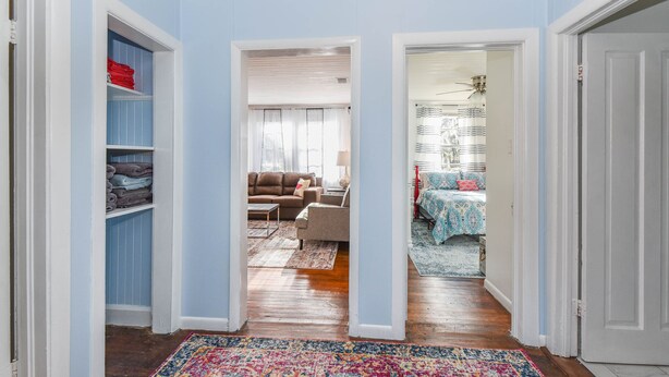 Un pasillo con suelo de madera, una alfombra colorida, una zona de estar con sofá y sillones, y un dormitorio con cama y cortinas.