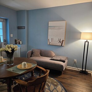 Ein Wohnzimmer mit einer Couch, einem Couchtisch, einem Esstisch mit Stühlen, einer Lampe und einem Kunstwerk mit Strandmotiv an der Wand.