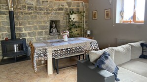 Un salon chaleureux avec un mur en pierre, une cheminée, une table à manger recouverte d’une nappe à motifs et un canapé gris agrémenté de coussins bleus.