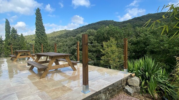 Une terrasse en pierre avec des tables de pique-nique et un muret en pierre, entourée de verdure et avec une chaîne de montagnes en toile de fond.