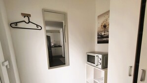 A narrow hallway with a white door, a black hanger on the wall, a microwave on a white cabinet, and a small black mat on the floor. - Vacation apartment Heide II (Walsrode)