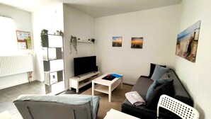 A modern living room with a flat-screen TV, a grey sofa, a white coffee table, and a small white side table. There are two framed pictures on the wall and a plant on a shelf. - Vacation apartment Heide II (Walsrode)