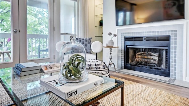 A living room with a fireplace, a glass coffee table with a book and a plant, and a view of the outdoors through large windows. - Stylish Uptown condo in Historic 4th Ward! (Charlotte)
