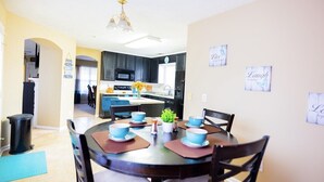 Un coin repas avec une table ronde dressée pour quatre personnes, des chaises en bois foncé et une cuisine en arrière-plan.