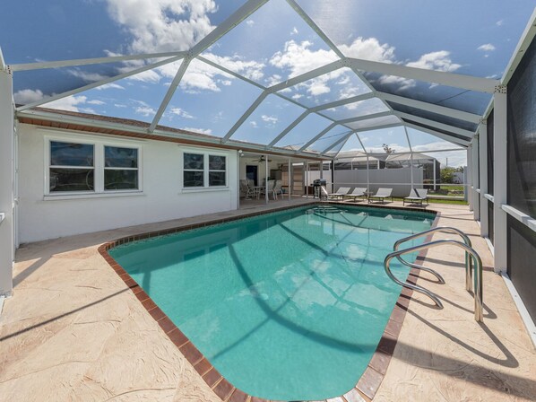 A covered swimming pool with a clear view of the sky, a patio area with chairs, and a building with windows.
