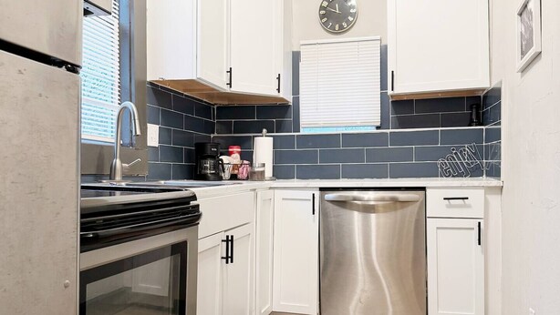 A compact kitchen with white cabinets, a stainless steel refrigerator, an oven, a dishwasher, and a clock on the wall. - Suite Home Indiana #2 (Evansville)