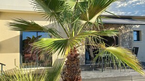 A modern building with a stone facade, large windows, and a palm tree in the foreground. - T3 apartement adjoining a luxury villa, 6 people, 70m2 (La Roquette-sur-Siagne)