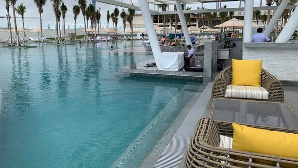 Un espace piscine doté de chaises longues et de parasols, de palmiers, ainsi que d’un espace de détente couvert avec un bar.