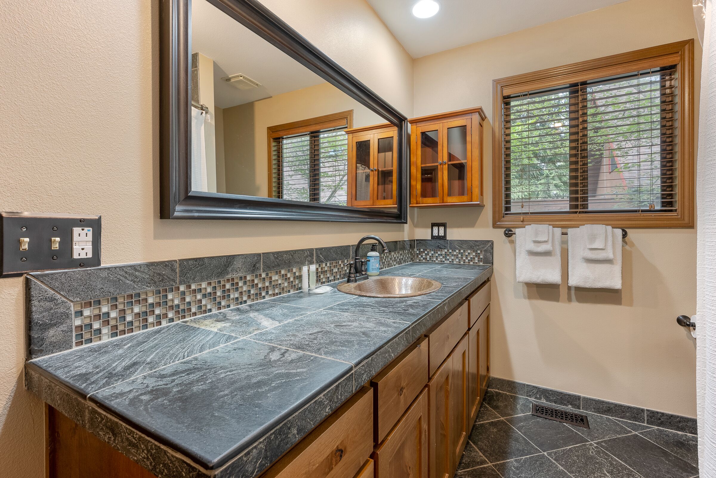 A bathroom with a granite countertop, a sink, wooden cabinets, a mirror, and a window with blinds.
