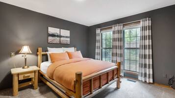 A bedroom with a wooden bed, a nightstand with a lamp, a window with curtains, and a wall-mounted picture.