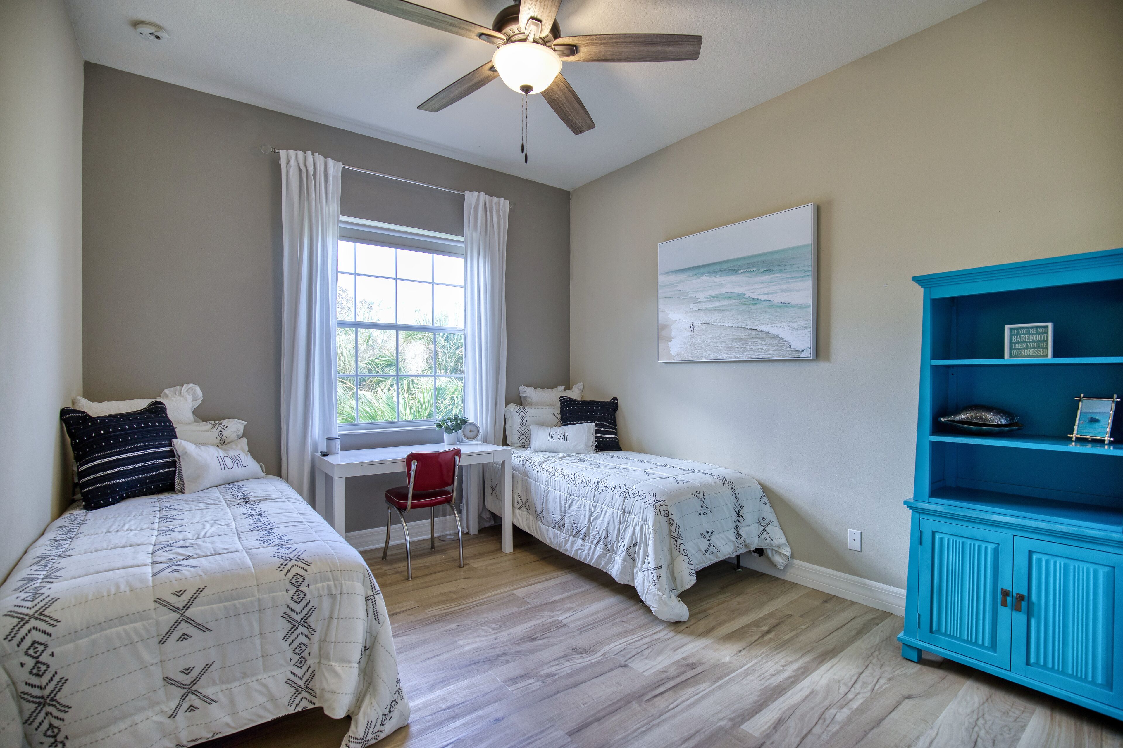 A bedroom with two beds, a desk, a chair, a ceiling fan, a window with curtains, a painting on the wall, and a blue cabinet.