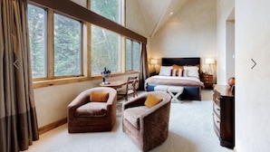 A spacious bedroom with a large bed, two armchairs, a desk, and a view of trees through large windows.
