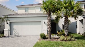 A two-story house with a white garage door, a brick driveway, and a well-maintained lawn with palm trees. - Beautiful home-private spa/pool 5BD 5.5 BA (Davenport)