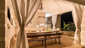 An outdoor dining area with a stone bench and a table set for two, surrounded by curtains and a stone wall.
