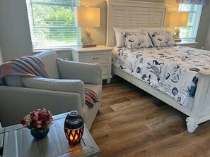 Une chambre à coucher comprenant un lit, une table de chevet, une lampe, une chaise et une table sur laquelle est posé un vase de fleurs.