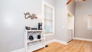 A modern kitchen with a white console table holding a coffee maker and a blender, a wall-mounted 'Coffee' sign, and a window with blinds.
