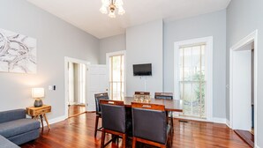 A spacious dining area with a wooden table and chairs, a television mounted on the wall, and a large window with blinds.