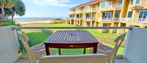 Un balcón con una mesa de madera y sillas blancas, con vistas a una playa y un edificio con balcones.