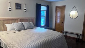 Ein Schlafzimmer mit einem Bett, einem Holz-Kopfteil, einem Deckenventilator, einem Fenster mit blauen Vorhängen, einer Tür und Wandleuchten.