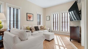 A spacious living room with a white sectional sofa, a coffee table, and a TV on a stand. There are large windows with blinds, a lamp on a side table, and a framed picture on the wall.