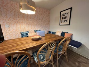 Een eetgedeelte met een houten tafel, rieten stoelen, een laptop en een muur met een 'PRAY FOR SURF' bord.