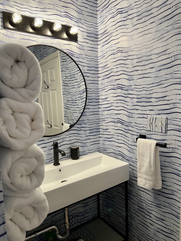 Un baño con un lavamanos blanco, un espejo redondo con luces arriba, un perchero para toallas y un papel tapiz decorativo.