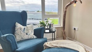 Un cuarto acogedor con un sillón azul y un reposapiés, una mesa con una planta y un hervidor, y vista al mar a través de un amplio ventanal.