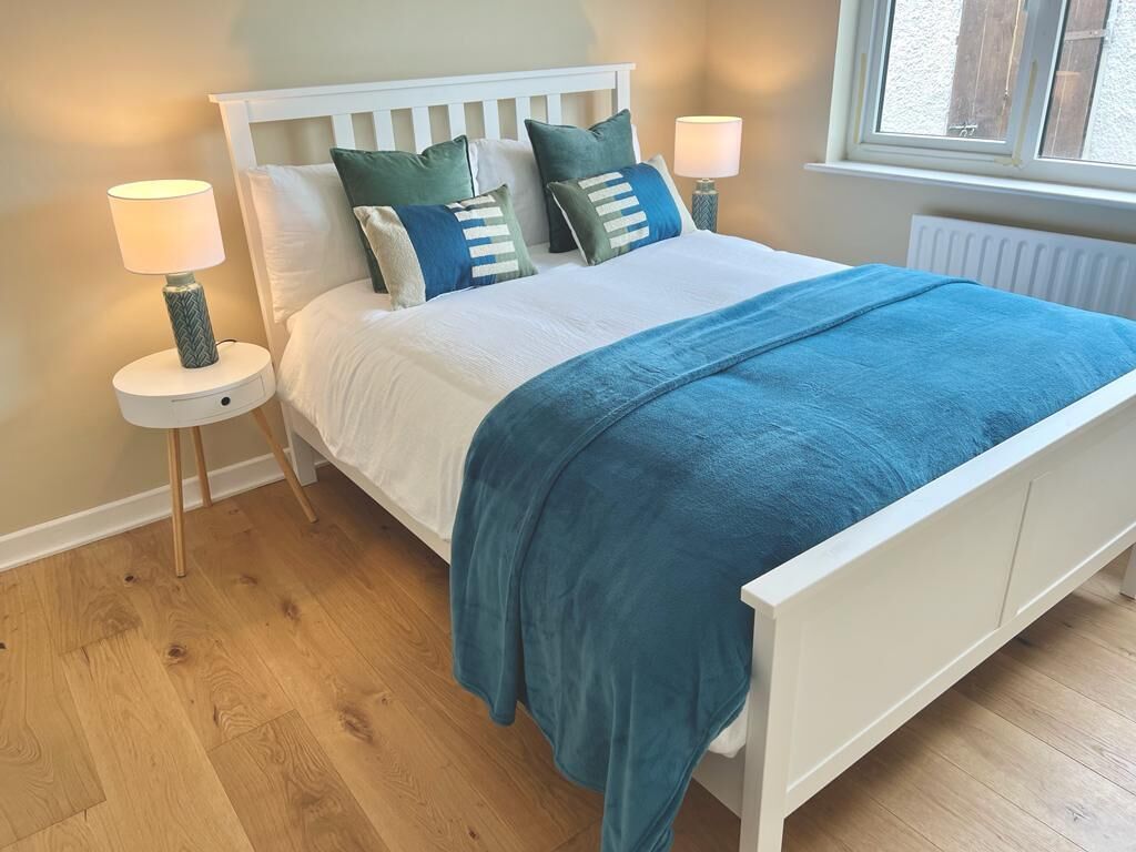 A neatly made bed with a white headboard, blue bedding, and multiple pillows. There are two bedside tables with lamps and a window with a view of the outside.