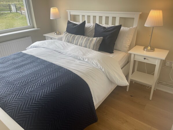 A neatly made bed with a white headboard and a dark blue chevron-patterned bedspread. There are two bedside tables with lamps, one on each side of the bed. - Luxury Donegal Beach Cottage at Dooey Beach, Dungloe (Lettermacaward)