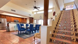 A spacious living area with a tiled floor, a dining area with blue chairs, a kitchen with wooden cabinets, and a decorative staircase. - Spectacular Views, Heated Pool, & Outdoor BBQ (Tucson)