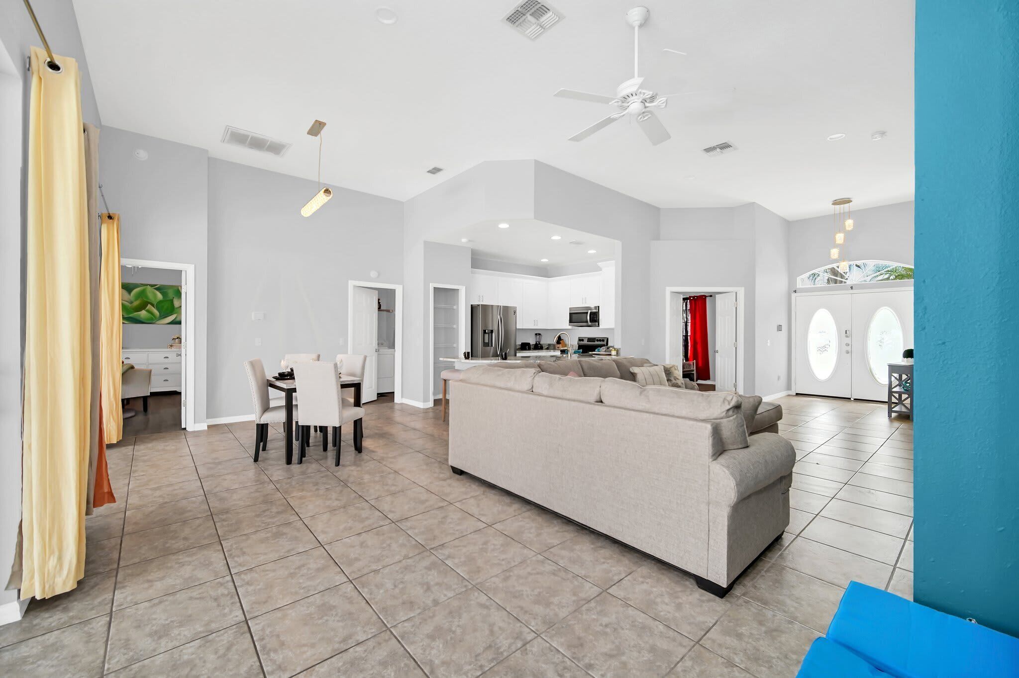 A spacious living area with a sofa, dining table, and chairs, a kitchen in the background, and a ceiling fan.
