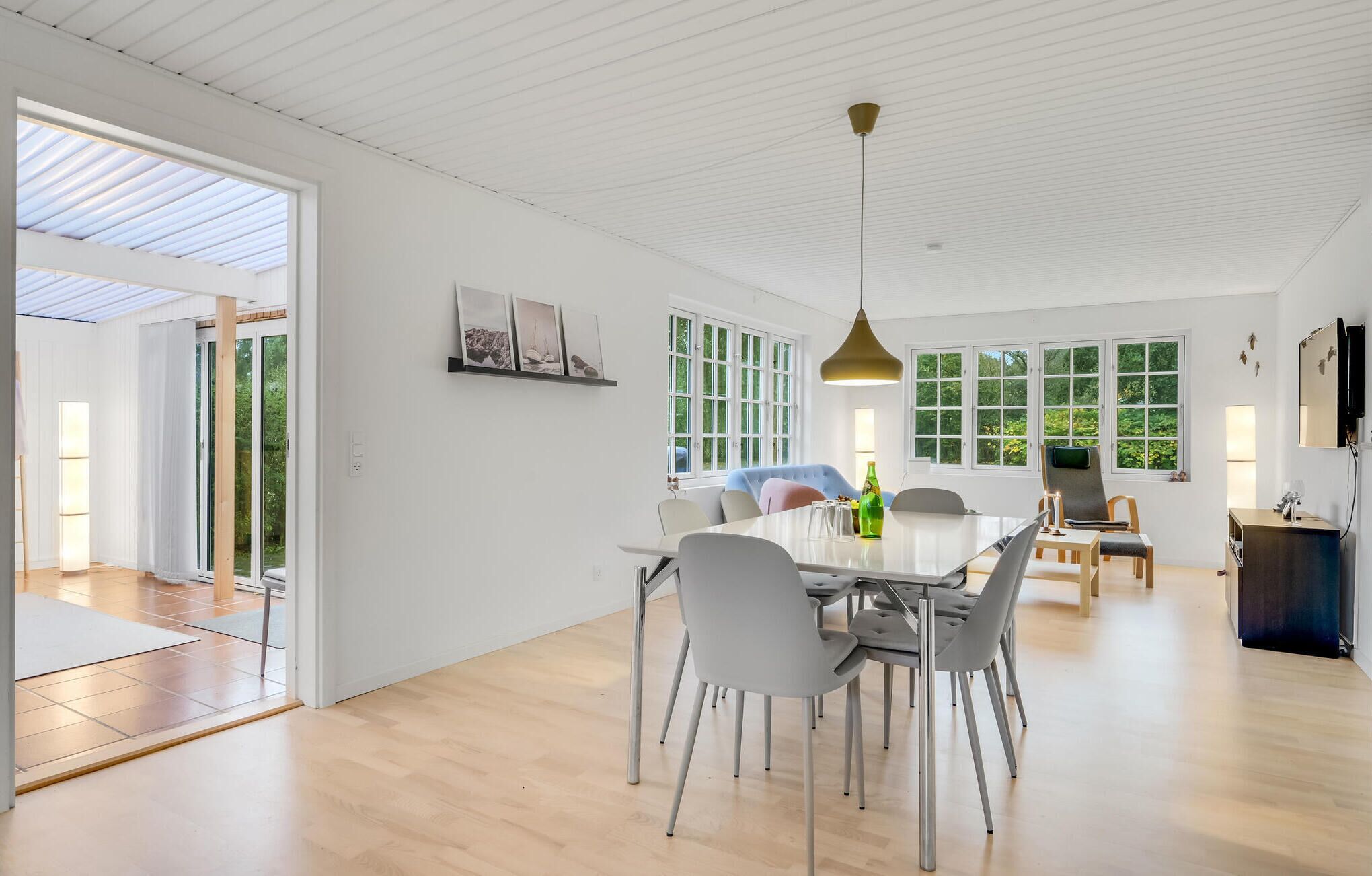 A spacious living area with a dining table, chairs, and a sofa. There are large windows providing natural light and views of greenery.