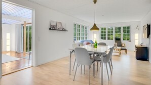 Amplia sala de estar con mesa de comedor, sillas y sofá. Grandes ventanales ofrecen luz natural y vistas al verdor.