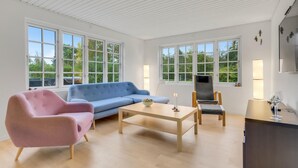 Una sala de estar moderna con un sillón rosa, un sofá azul, una mesa de centro de madera y un sillón gris. Se observan ventanas grandes con marcos blancos, un televisor montado en la pared y una mesita con una vela y un vaso.