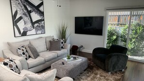 A modern living room with a grey sofa, a black armchair, a coffee table, and a large TV. There is a window with a view of greenery outside. - Sweet cottage home close to beaches, airport and other attractions (San Diego)
