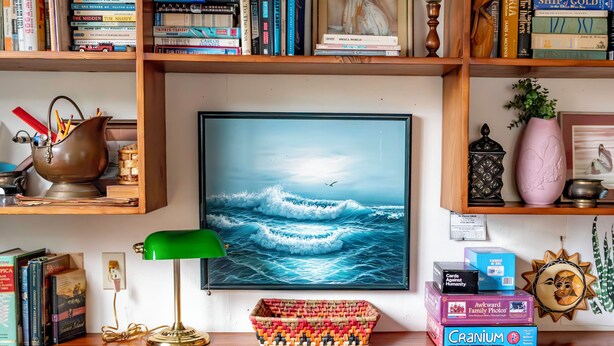 Une bibliothèque remplie de divers livres, un tableau encadré représentant l’océan, une lampe de bureau et un panier en osier.