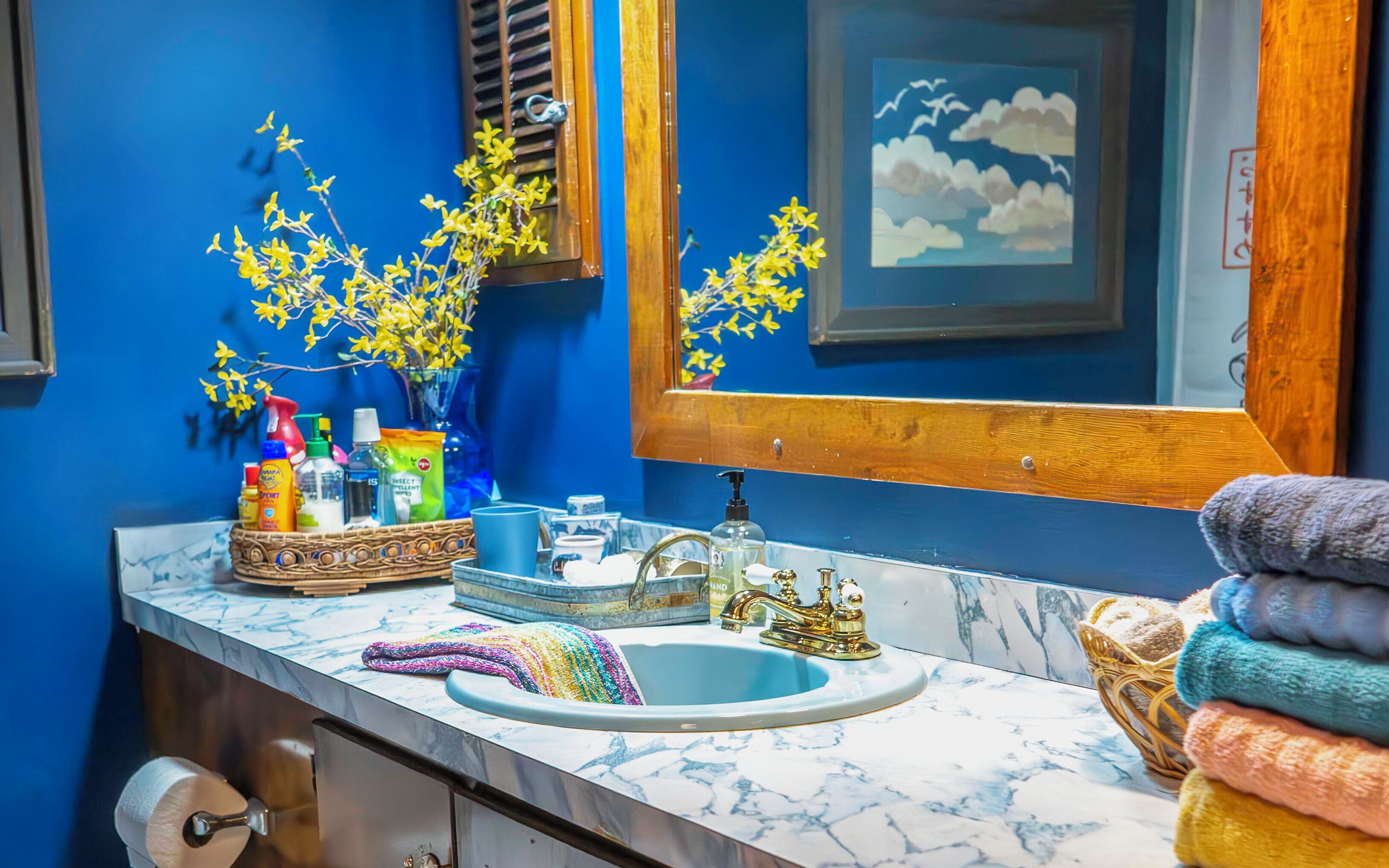 Un baño con pared azul, un lavamanos de mármol y un espejo. Hay artículos de aseo en la encimera, un perchero y un cuadro en la pared.
