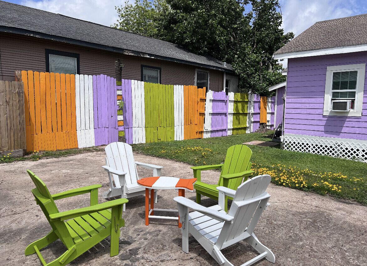 Een terras met Adirondack-stoelen en een kleine tafel, een kleurrijke houten omheining, en een huis met een paarse gevel.