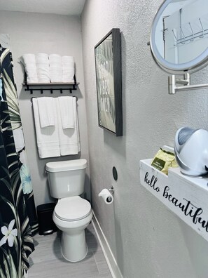 A bathroom with a toilet, a towel rack with folded towels, a framed picture on the wall, a toilet paper holder, and a shelf with a facial cleansing wipes package.