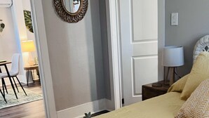 A bedroom with a bed, a bedside table, a lamp, a mirror, and a door leading to another room.