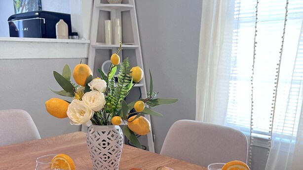 A dining table set with plates, cutlery, a loaf of bread, glasses of water, and a vase with flowers and lemons.
