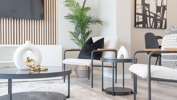 Un salon moderne avec un canapé, une table basse et des tables d’appoint. On y trouve une grande plante, un tableau accroché au mur et un téléviseur fixé au-dessus du canapé.