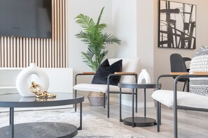 Un salon moderne avec un canapé, une table basse et des tables d’appoint. On y trouve une grande plante, un tableau accroché au mur et un téléviseur fixé au-dessus du canapé.