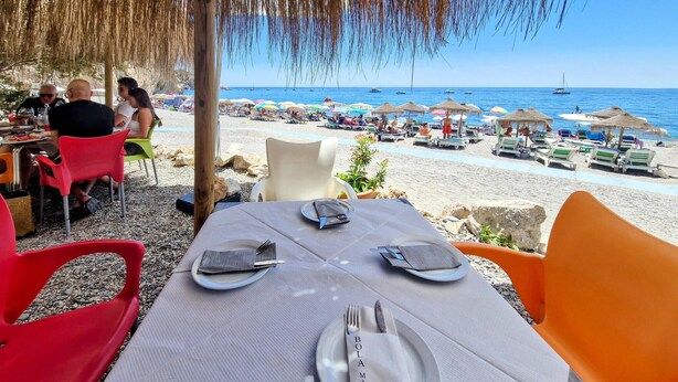 En restaurang vid stranden med uteservering, utsikt över havet och ett dukat bord med tallrikar och bestick.